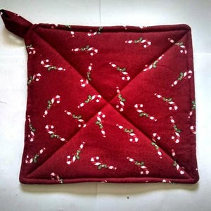 May include: A square, quilted potholder in a deep red fabric. The potholder is decorated with a repeating pattern of white and green candy canes. It has a small loop for hanging.