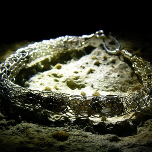 Puede incluir: Un collar de cadena de plata con un diseño tejido y cuentas de plata. El collar está sobre una superficie texturizada.