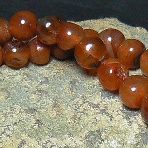 May include: A strand of carnelian beads in shades of orange and brown. The beads are round and smooth, and they are strung on a clear thread.