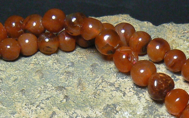 May include: A strand of carnelian beads in shades of orange and brown. The beads are round and smooth, and they are strung on a clear thread.