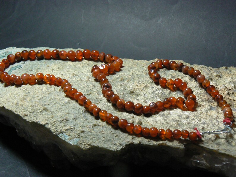 May include: A long strand of carnelian beads, with a shorter strand of carnelian beads attached to it. The beads are a deep orange-red color and are strung on a silver cord.