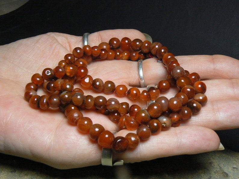 May include: A long strand of carnelian beads, with a reddish-brown color, is displayed on a hand. The beads are round and smooth, and they are strung together on a clear thread.