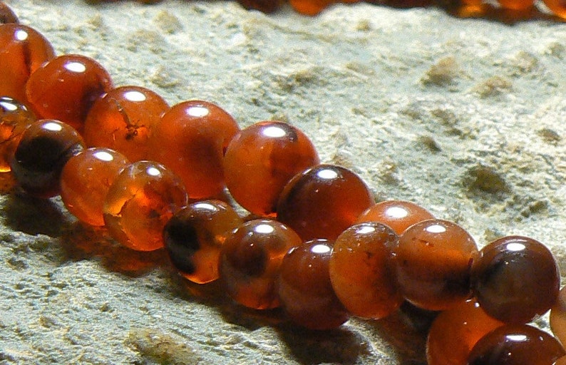 May include: A strand of carnelian beads, each bead is a deep orange-red color with a slight translucence. The beads are round and smooth, and they are strung together on a clear thread.