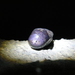 Puede incluir: Una piedra preciosa de color púrpura oscuro con una superficie lisa y pulida. La piedra preciosa descansa sobre una superficie rugosa y gris.