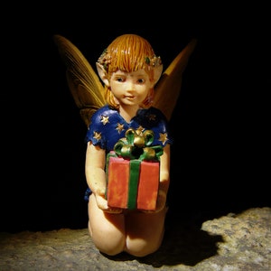 May include: A fairy figurine with wings, wearing a blue and gold dress, holding a red and green gift box. The figurine is posed kneeling on a brown surface.