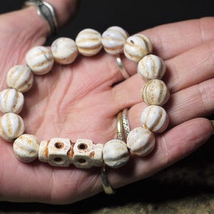 May include: A white and brown beaded bracelet with a decorative, carved bone charm. The beads are round and have a ribbed texture.
