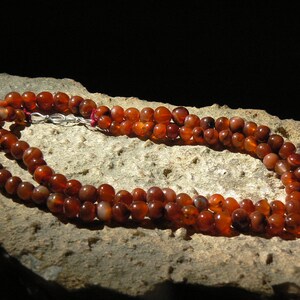 May include: A double strand necklace made of carnelian beads. The beads are a deep orange-red color and have a natural, polished finish. The necklace is displayed on a rough, brown surface.