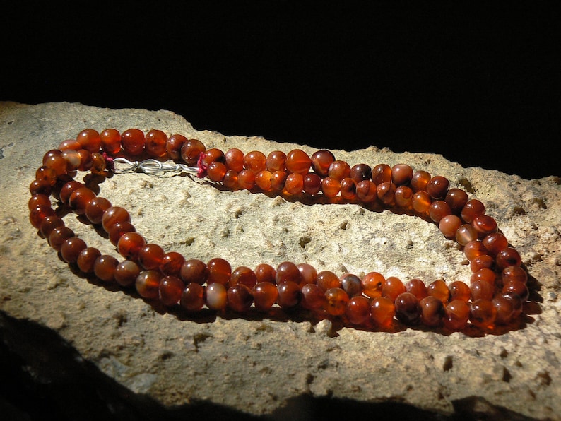 May include: A double strand necklace made of carnelian beads. The beads are a deep orange-red color and have a natural, polished finish. The necklace is displayed on a rough, brown surface.