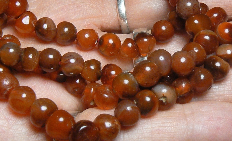 May include: A strand of carnelian beads, featuring a rich, reddish-brown color with subtle variations in tone. The beads are smooth and polished, creating a lustrous finish.