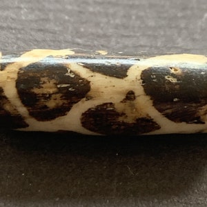 Peut inclure: Une perle cylindrique avec un motif marron foncé et crème. La perle a une surface lisse et polie et une forme légèrement arrondie. Le motif semble organique et irrégulier, avec des formes marron foncé sur un fond crème. La perle est probablement en bois.