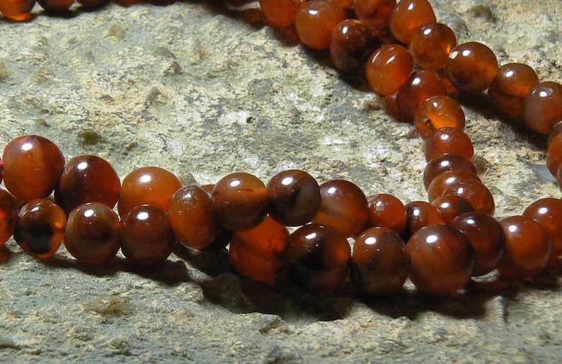 May include: A strand of carnelian beads, a type of chalcedony, in shades of orange and brown. The beads are round and smooth, and they are strung on a cord.