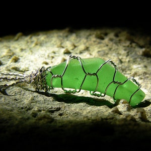 May include: A green sea glass pendant wrapped in silver wire, creating a delicate and intricate design. The pendant is lying on a rough, textured surface.