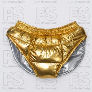 Puede incluir: Pantalones cortos acolchados de color dorado metálico con acabado brillante y cintura elástica. La prenda presenta un forro plateado visible en las aberturas de las piernas y una construcción acolchada con costuras en cuadrícula.