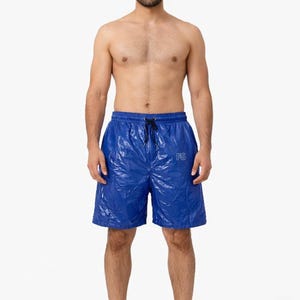 May include: Bright blue swim shorts with a drawstring waist and the letters "PS" printed on the leg. The shorts have a shiny, crinkled texture. The model is standing in front of a white background.