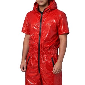 May include: A shiny red jumpsuit with a hood and short sleeves. The jumpsuit has a black zip down the front and black zips on the pockets. The model is wearing a black shirt underneath.