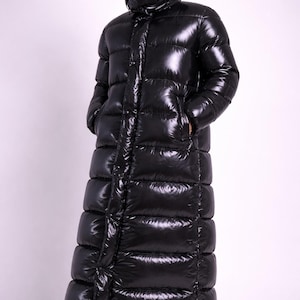 May include: A full-length, glossy black puffer coat with a hood. The coat has a high collar and a quilted design. It features a zipper closure and pockets. The person is wearing blue jeans and black shoes.