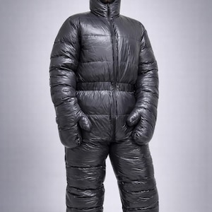 Puede incluir: Mono acolchado gris de cuerpo entero con capucha y mitones adjuntos. El traje tiene una cremallera frontal y una cintura ceñida. El material tiene una apariencia brillante y acolchada. Zapatos grises a juego.
