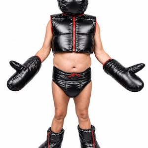 Op de afbeelding: Een zwarte gewatteerde puffer-set bestaande uit een mouwloos vest met ritssluiting, een capuchonmasker met ronde oogopeningen, een slip met hoge taille en trekkoord, oversized wanten en enkellaarsjes, allemaal voorzien van rode accenten.