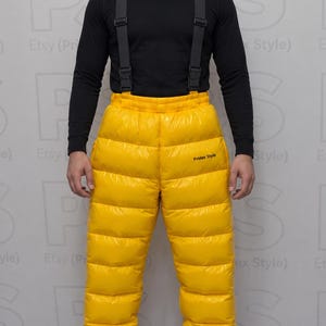 Puede incluir: Una camiseta negra de manga larga combinada con pantalones de plumón acolchados de color amarillo brillante con tirantes negros. Los pantalones tienen cintura alta, construcción de paneles horizontales y el texto Pridex Style impreso en negro.