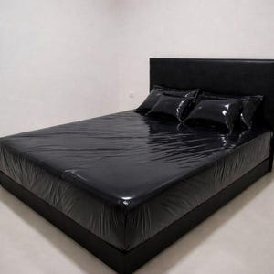 Op de afbeelding: Een zwart bed met een hoofdbord en drie zwarte kussens. Het bed is bedekt met een glanzend, zwart laken. De kamer heeft witte muren en een lichtgekleurde vloer.