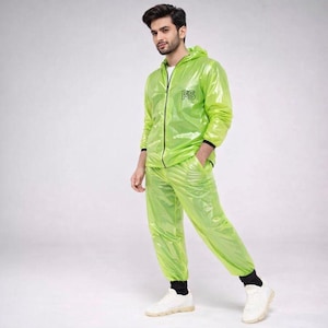 Könnte beinhalten: Ein neongrüner Regenschutzanzug, bestehend aus einer Kapuzenjacke mit Reißverschluss und passender Hose. Auf der Jacke sind die Buchstaben "PS" auf der Brust aufgedruckt. Der Anzug wird mit weißen Turnschuhen und schwarzen Socken getragen. Das Material scheint ein glänzendes, wasserdichtes Gewebe zu sein.