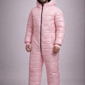May include: A pale pink, padded all-in-one snowsuit with a hood and a black zip. The one-piece garment has a shiny, puffy appearance and is designed for cold weather. The suit covers the entire body, including the feet.