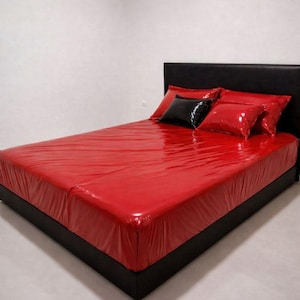 Può includere: Un letto con testiera e struttura neri, coperto da un lenzuolo rosso lucido. Due cuscini rossi e un cuscino nero sono sul letto, creando un contrasto audace. La stanza ha pareti bianche e pavimento chiaro.