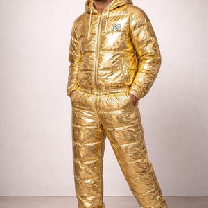 Puede incluir: Conjunto de chaqueta y pantalón acolchados dorados. La chaqueta tiene capucha y cierre de cremallera. El pantalón tiene cintura elástica. El conjunto se lleva con zapatillas blancas. La chaqueta tiene las letras "PS".