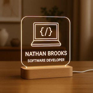 Può includere: Lampada da scrivania a LED personalizzata con base in legno. Il pannello in acrilico trasparente presenta un'icona di laptop con parentesi graffe, il nome "NATHAN BROOKS" e il titolo "SOFTWARE DEVELOPER" in bianco. La lampada emette una luce calda e soffusa.
