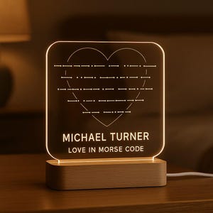 May include: A personalised night light featuring a heart in Morse code. The light is engraved with the name "MICHAEL TURNER" and the phrase "LOVE IN MORSE CODE". It rests on a wooden base and emits a warm glow.