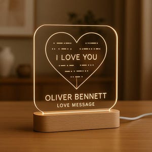 May include: An illuminated acrylic night light in the shape of a heart, featuring the text "I LOVE YOU", a name, "OLIVER BENNETT", and "LOVE MESSAGE". It is mounted on a wooden base and connected to a power cord.