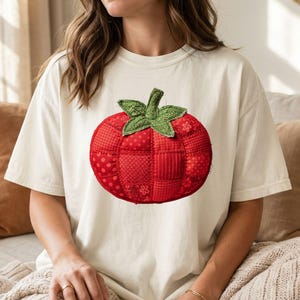May include: Cream-colored t-shirt featuring a large, embroidered tomato design. The tomato is red with a patchwork pattern and green leaves. The shirt is a relaxed fit, suitable for casual wear.