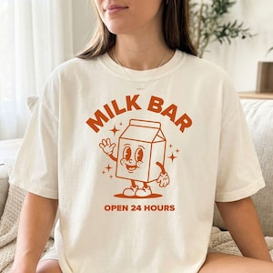 以下が含まれることがあります： レトロスタイルのグラフィックが特徴のクリーム色のTシャツ。デザインは、オレンジ色の文字で「MILK BAR」と書かれた笑顔の牛乳パックのキャラクターと、「OPEN 24 HOURS」が特徴です。このシャツはカジュアルな半袖スタイルです。