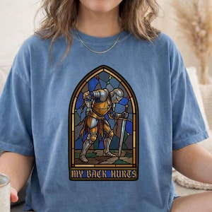 Może przedstawiać: Niebieski t-shirt z krótkim rękawem z grafiką rycerza w zbroi, który pochyla się, trzymając miecz, wkomponowany w witrażowy wzór. Napis "MY BACK HURTS" jest wydrukowany pogrubioną, ciemną czcionką w dolnej części grafiki na koszulce.