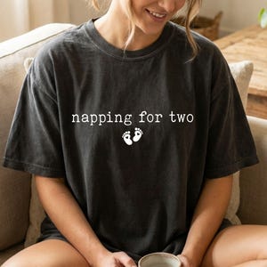 May include: Dark gray t-shirt with the text "napping for two" and two small white footprints. The shirt is a relaxed fit and appears to be made of a soft material. The shirt is a casual style.