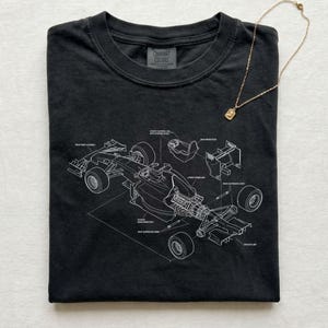 May include: Black t-shirt with a white diagram of a race car, featuring labeled parts. A gold necklace with a square pendant rests on the shirt. The shirt has a crew neck and the brand name "Comfort Colors" is visible.