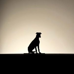 以下が含まれることがあります： 座っている犬の黒いシルエット。犬は長方形の台座の上にあり、背景は明るいです。犬の耳はピンと立ち、尾は下がっています。画像はシンプルでエレガントです。