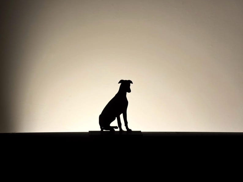 以下が含まれることがあります： 座っている犬の黒いシルエット。犬は長方形の台座の上にあり、背景は明るいです。犬の耳はピンと立ち、尾は下がっています。画像はシンプルでエレガントです。