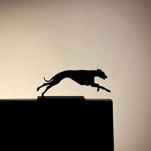 以下が含まれることがあります： 暗い長方形の土台の上に、走っている犬、おそらくグレイハウンドの黒いシルエットがあります。犬は、脚を伸ばして、速度と動きの感覚を伝える、途中で捉えられています。背景は明るいベージュのグラデーションです。