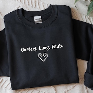 May include: Black sweatshirt with the text "Ua Neej. Luag. Hlub." in white, along with a white stitched heart design. The sweatshirt is made of a soft material and has a crew neck.