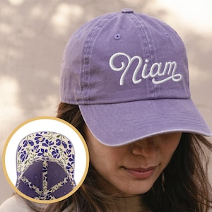 May include: A purple baseball cap with the name "Niam" embroidered in white script. The cap has a curved brim and a floral pattern on the underside. The hat is made of a soft, worn material.