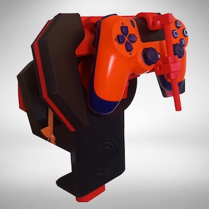May include: A black and orange gaming controller mount featuring a structured, angular design with a prominent vertical support arm. The mount is colored in matte black with bright orange accents and holds a matching orange and blue game controller.