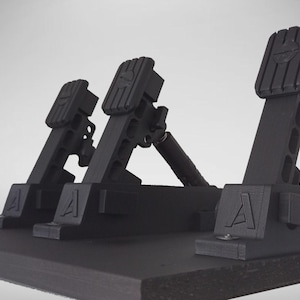May include: A set of three black racing simulator pedals mounted on a flat base. Each pedal features a vertical arm with a textured foot plate, a spring mechanism, and a prominent letter A embossed on the base. The design is industrial and matte.