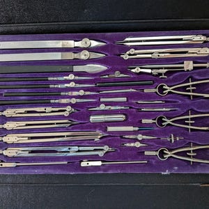 May include: A vintage drafting instrument set housed in a black case with a purple velvet interior. The collection includes various silver-toned metal compasses, dividers, ruling pens, and extension bars arranged in custom-molded slots.