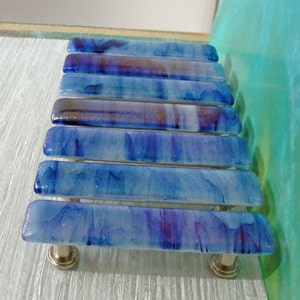 May include: A set of eight blue and purple glass drawer pulls with a swirled pattern. The pulls are mounted on a white surface.