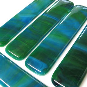 May include: Close-up of several rectangular glass pieces with a glossy finish. The glass features a blend of blue and green hues, creating a marbled effect. The pieces are arranged on a white surface, showcasing their vibrant colors.
