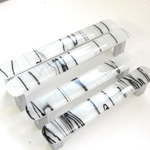 May include: Four white glass cabinet pulls with black abstract lines. The pulls are rectangular and have a rounded edge on one end.