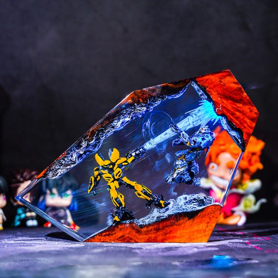 A hand-poured resin diorama lamp that freezes Megatron and Bumblebee mid-battle inside a glowing wood-base night light — genuinely one-of-a-kind nerdy craft art.