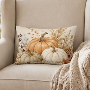 May include: A rectangular throw pillow featuring a printed design of orange and white pumpkins surrounded by dried wheat and autumn foliage. The pillow is placed on a cream-colored upholstered wingback chair with a textured beige knit throw blanket.