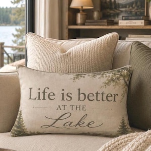 May include: A rectangular beige throw pillow featuring the printed text Life is better AT THE Lake in dark brown lettering. The design includes illustrations of evergreen trees at the bottom corners and subtle tree foliage along the top edge.